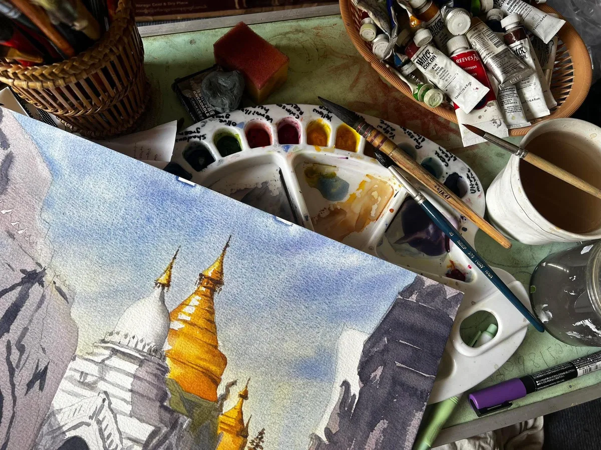 Watercolor painting of a pagoda in progress, surrounded by paints and brushes