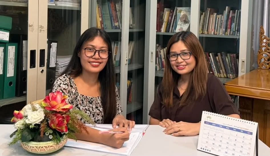 Two young women seated at a desk with bookshelves behind them